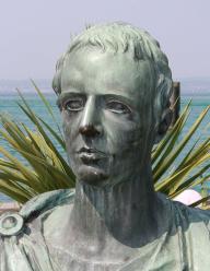 A statue bust of a man, the ocean behind him, green leaves
