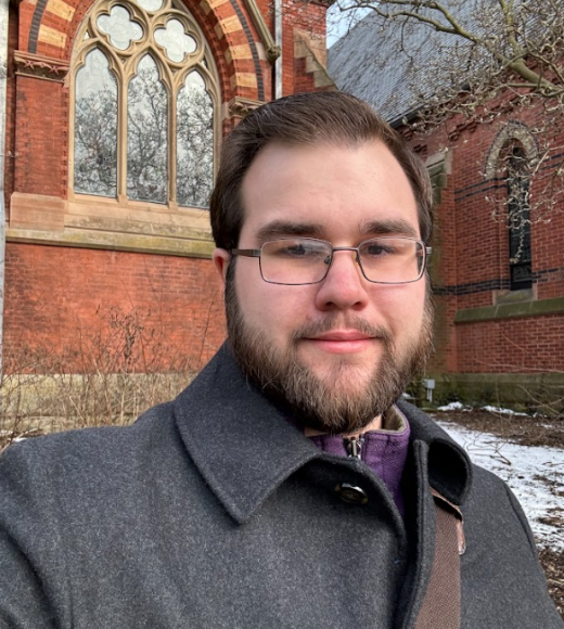 Terek stands in front of a brick building with an ornate arched window.