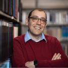 A photo of a man with glasses and red seater leaning against a bookshelf.